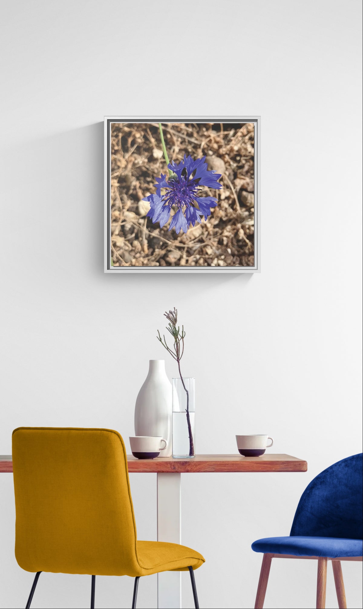 Framed wall art of a blue cornflower above a dining table with chairs and vases