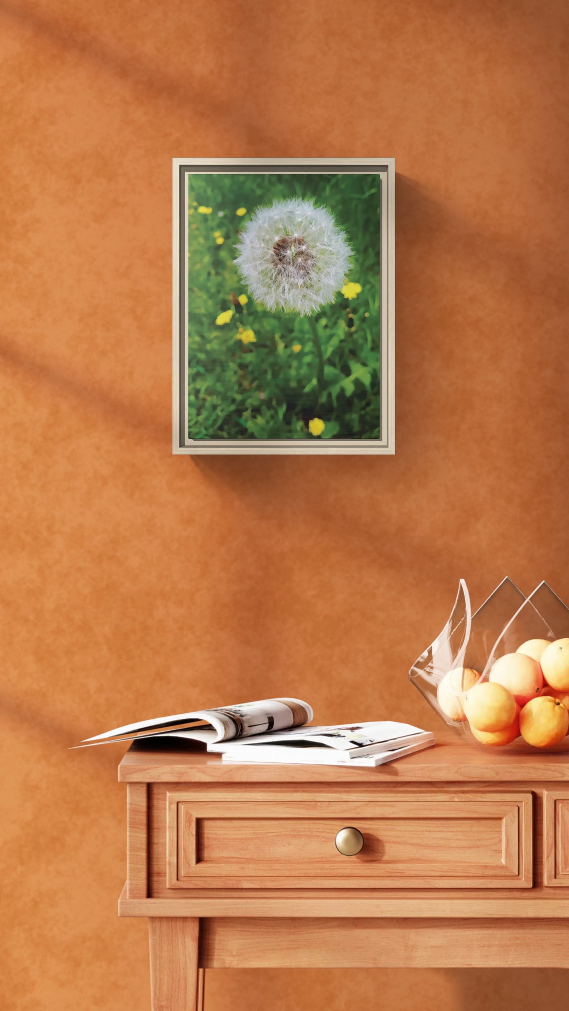 Framed wall art of a dandelion seed head on a textured orange wall above a wooden console table with fruit
