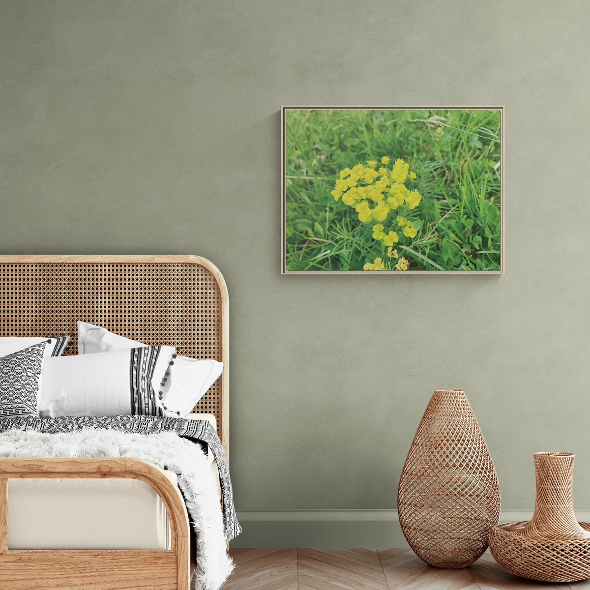 A framed wall art print depicting a cluster of bright yellow wildflowers in a green grassy field, displayed on a textured sage green wall above a wooden bed frame in a styled bedroom setting.