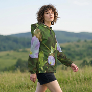 A woman wearing a green hooded jacket with large purple and white flowers printed on the sleeves, standing in a grassy field with hills in the background.