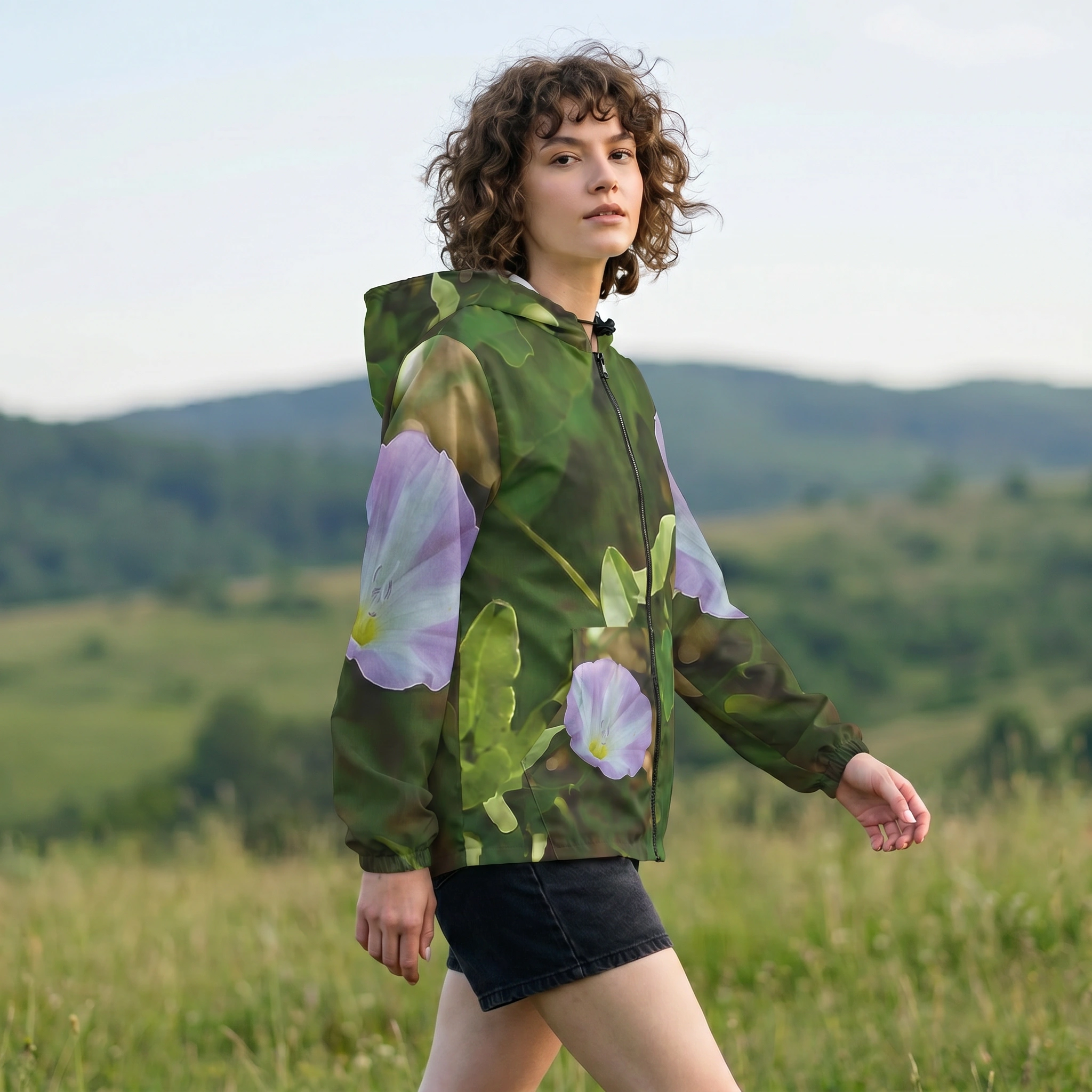 A woman wearing a green hooded jacket with large purple and white flowers printed on the sleeves, standing in a grassy field with hills in the background.
