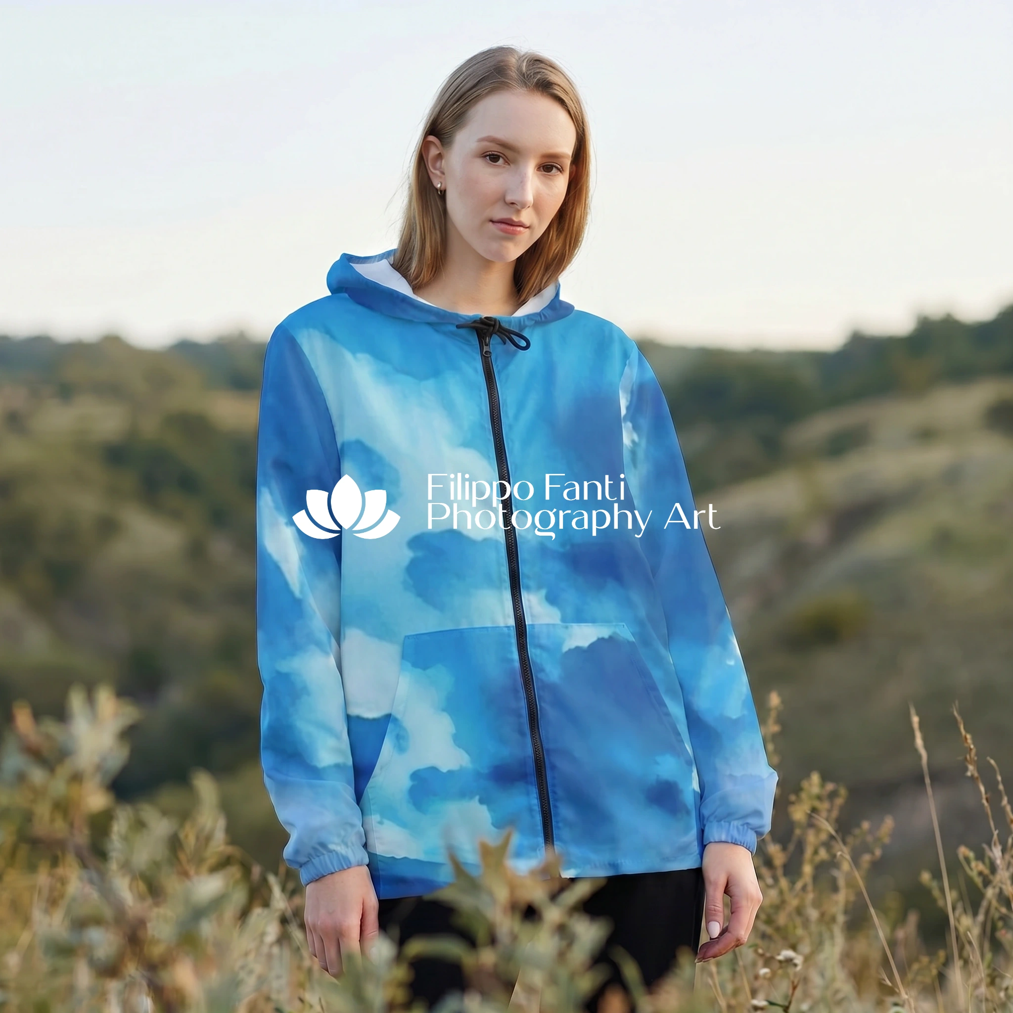 A woman standing outdoors in a grassy field wearing a blue hooded sweatshirt with a cloudy watercolor pattern. The image features a white watermark logo for 'Filippo Fanti Photography Art' on the front of the hoodie.