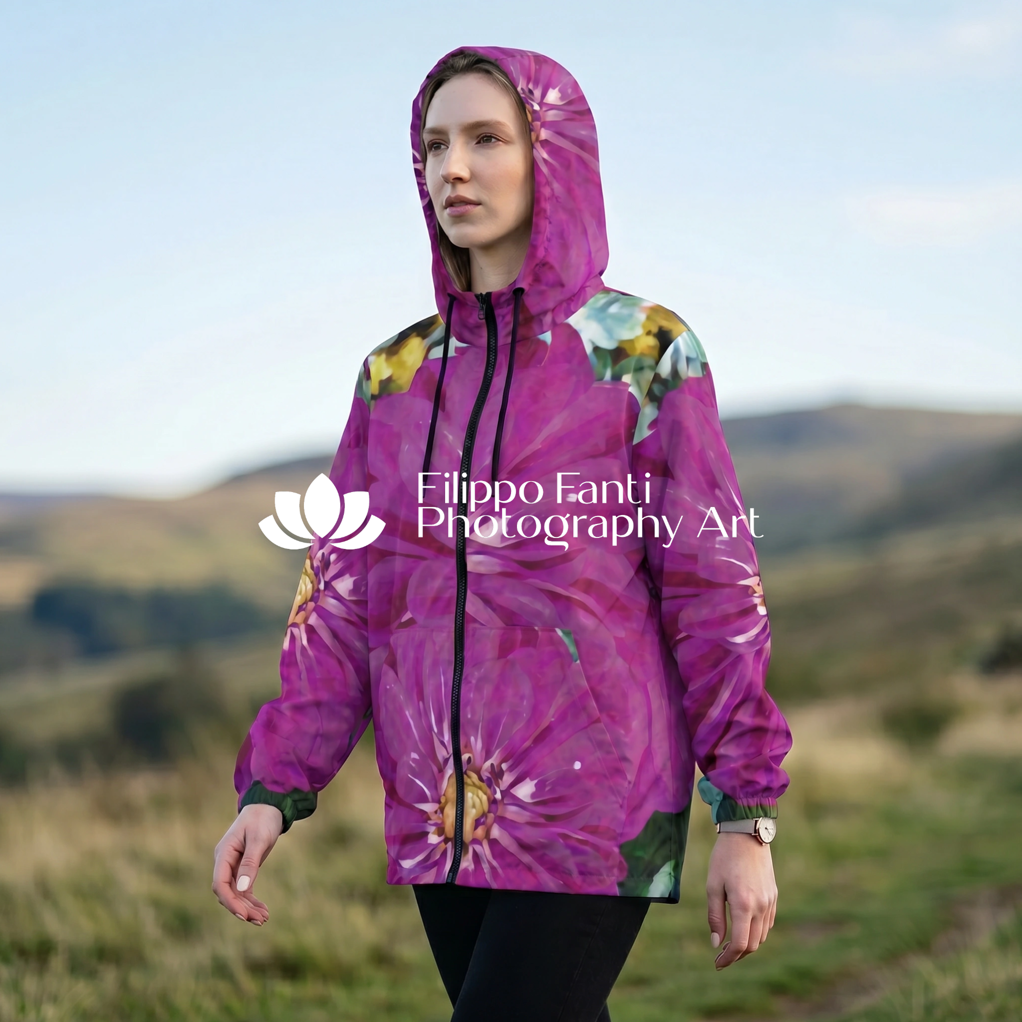 A woman wearing a hooded jacket with a large purple floral print stands outdoors in a grassy landscape. The jacket features a bold magenta and purple floral pattern with green and yellow accents.