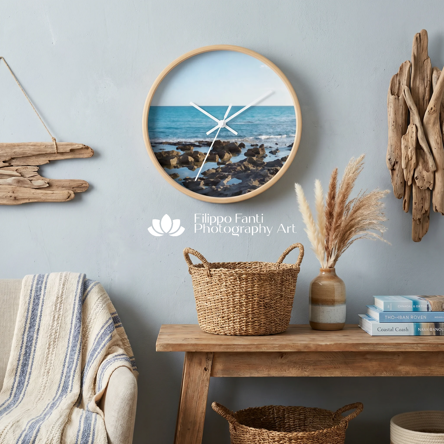 A round wall clock with a light wood frame hangs on a pale blue-gray wall above a wooden bench. The clock face shows a beach and ocean scene with white clock hands.