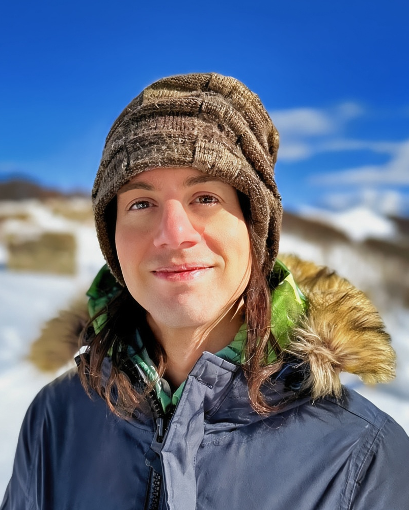 A man wearing a brown knit beanie and a navy blue winter parka with a fur-trimmed hood stands outdoors in a snowy landscape under a bright blue sky.