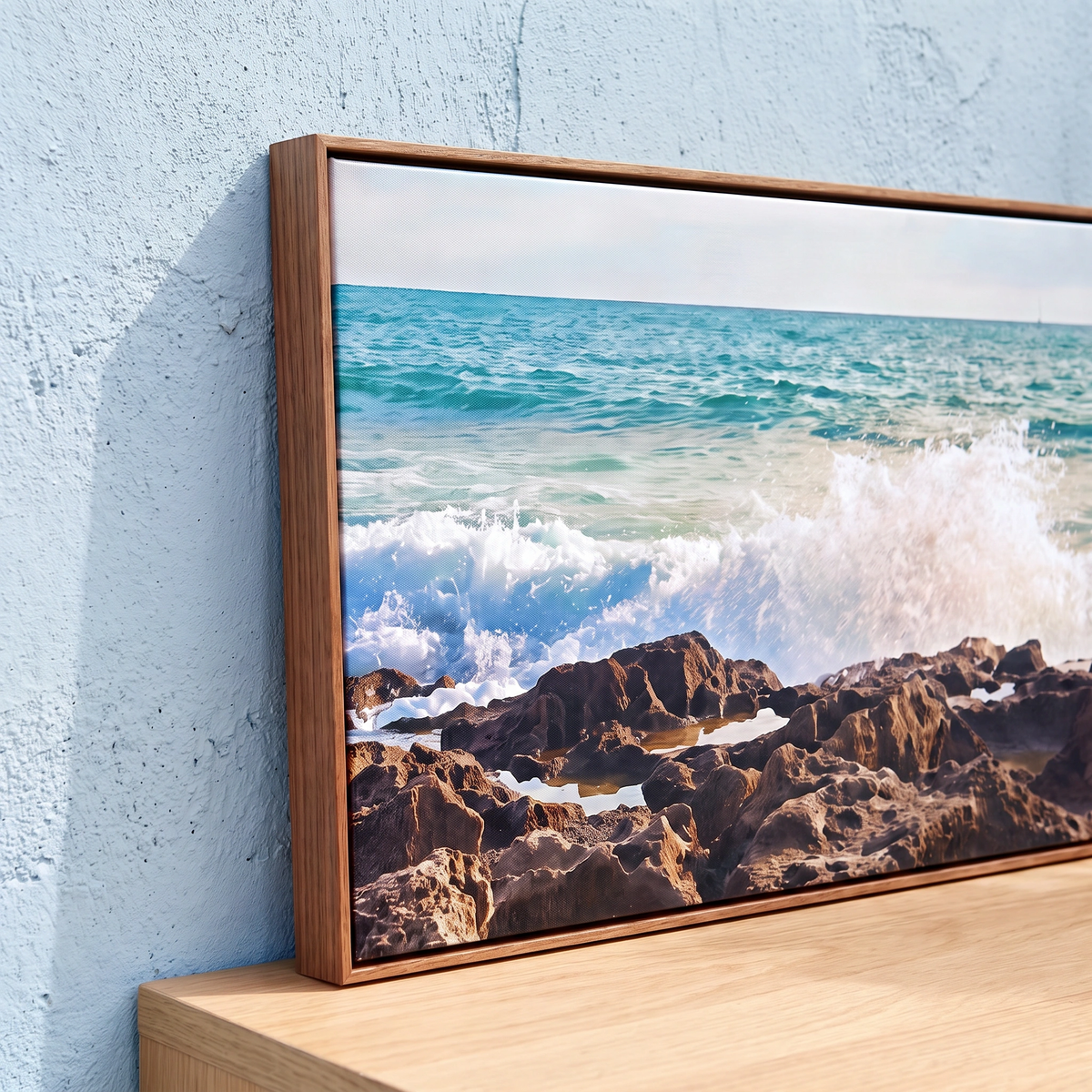 A framed horizontal canvas print of a rocky beach and turquoise ocean waves, leaning against a light blue textured wall on a light wood surface.