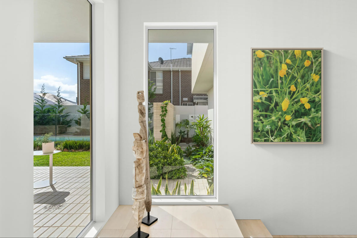 Modern interior with wooden sculptures and a framed yellow flower print on a white wall overlooking a garden