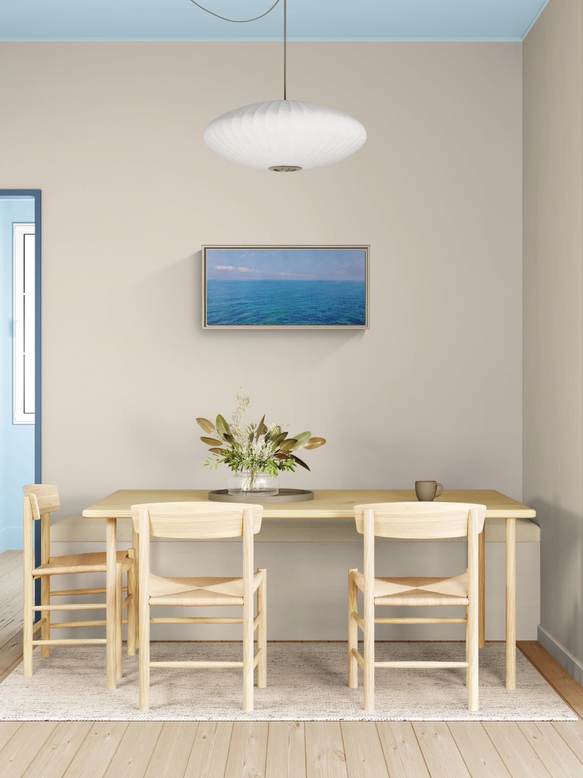 Framed ocean landscape art hanging above a wooden dining table with chairs and a vase of greenery