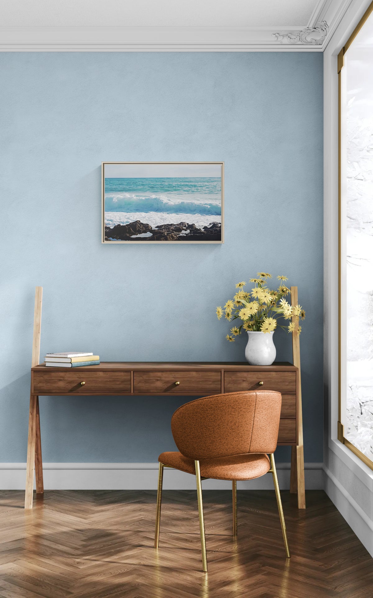 Framed ocean wave print hanging above a wooden desk with a chair and flowers