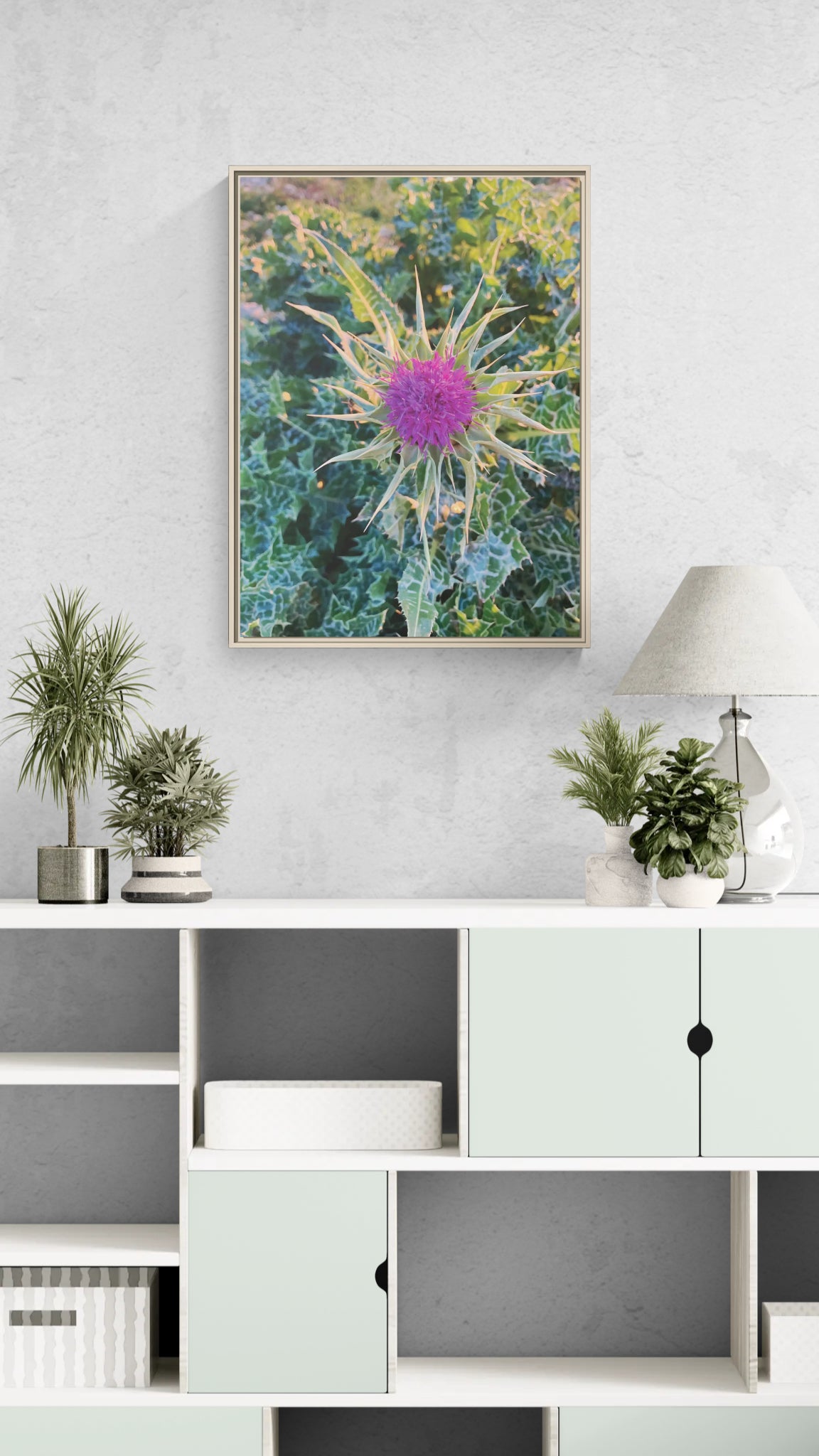 Framed wall art featuring a purple thistle flower on a white sideboard with potted plants and a lamp