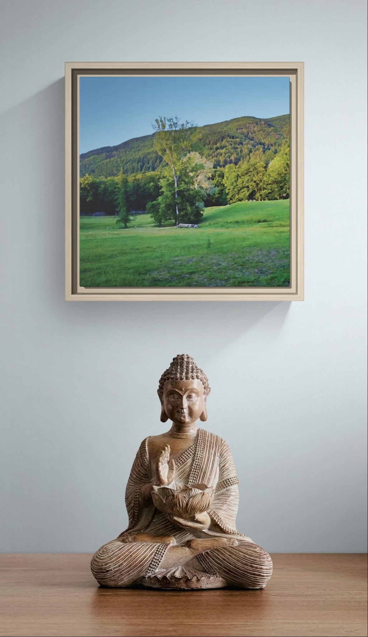 Framed landscape print of a green meadow and forest with a Buddha statue on a wooden surface below
